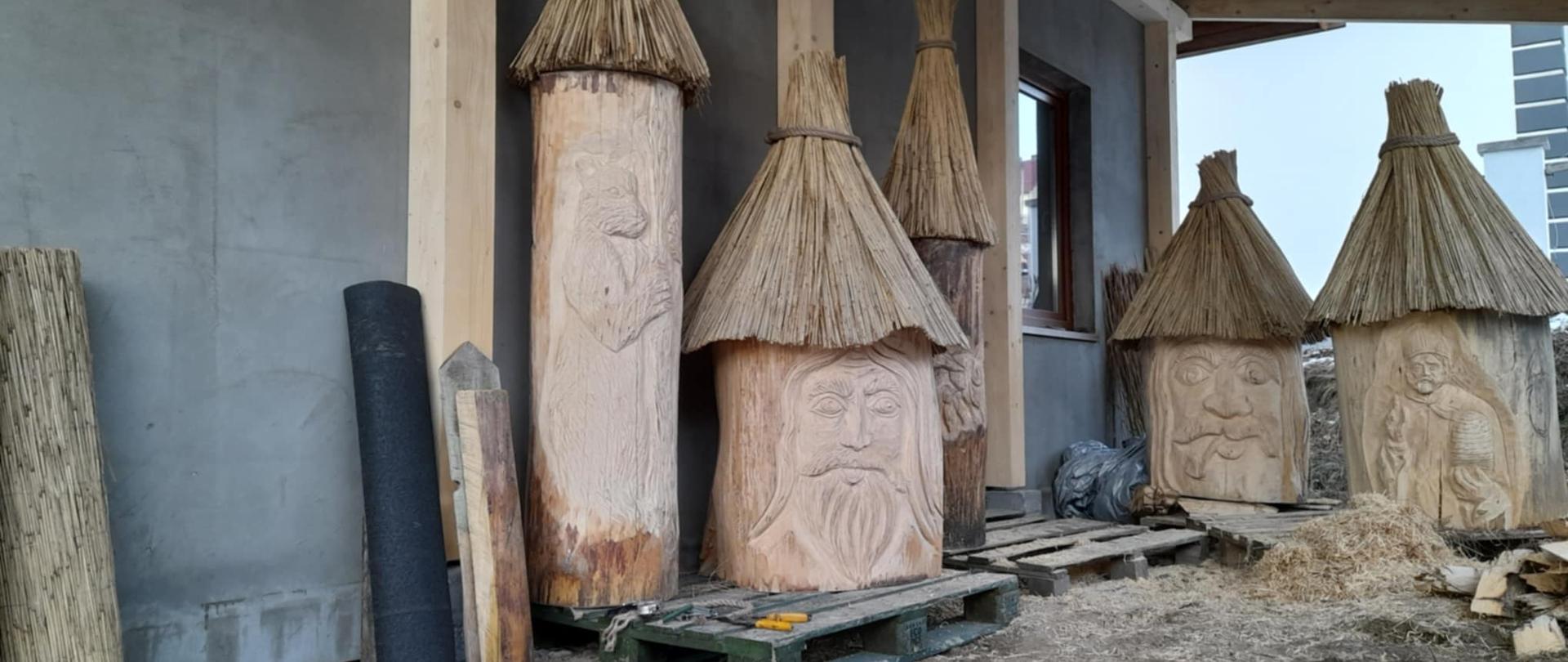 Pod dachem obok domu stoją rzeźby z drewna, przedstawiające ludzi oraz zwierzę.