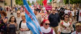 Parada w ramach Międzynarodowego Festiwalu Folklorystycznego Polka. Uczestnicy w strojach ludowych niosą flagi narodowe, maszerując ulicą miasta. Widać grupę w meksykańskich sombrero, niosącą flagę Meksyku. W tle tłum widzów i uczestników oraz zabudowa miejska.
