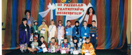 Grupa teatralna pozuje do wspólnego zdjęcia na tle ścianki z napisem III Przegląd Teatrzyków Dziecięcych