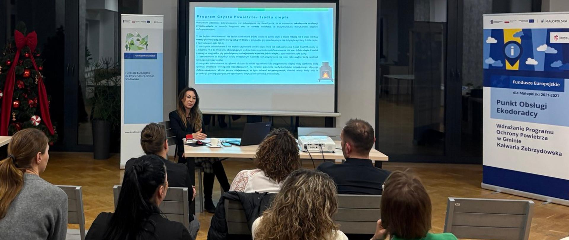 Mieszkańcy podczas spotkania z ekodoradcą siedzą na krzesłach, prowadząca przedstawia prezentację na ekranie, po bokach rollupy promujące