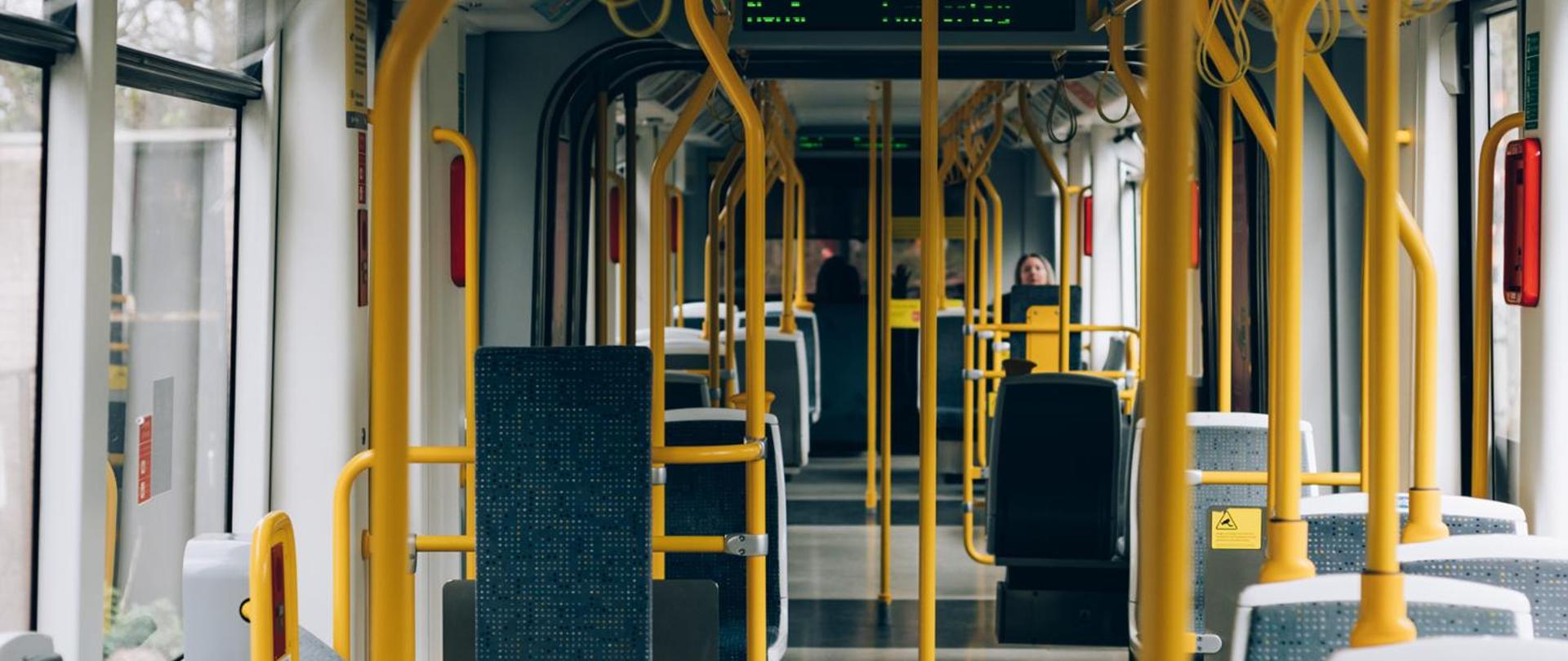 Wnętrze nowoczesnego tramwaju z żółtymi poręczami, rzędami siedzeń i szerokim przejściem po środku. W oddali widocznych jest kilku pasażerów.