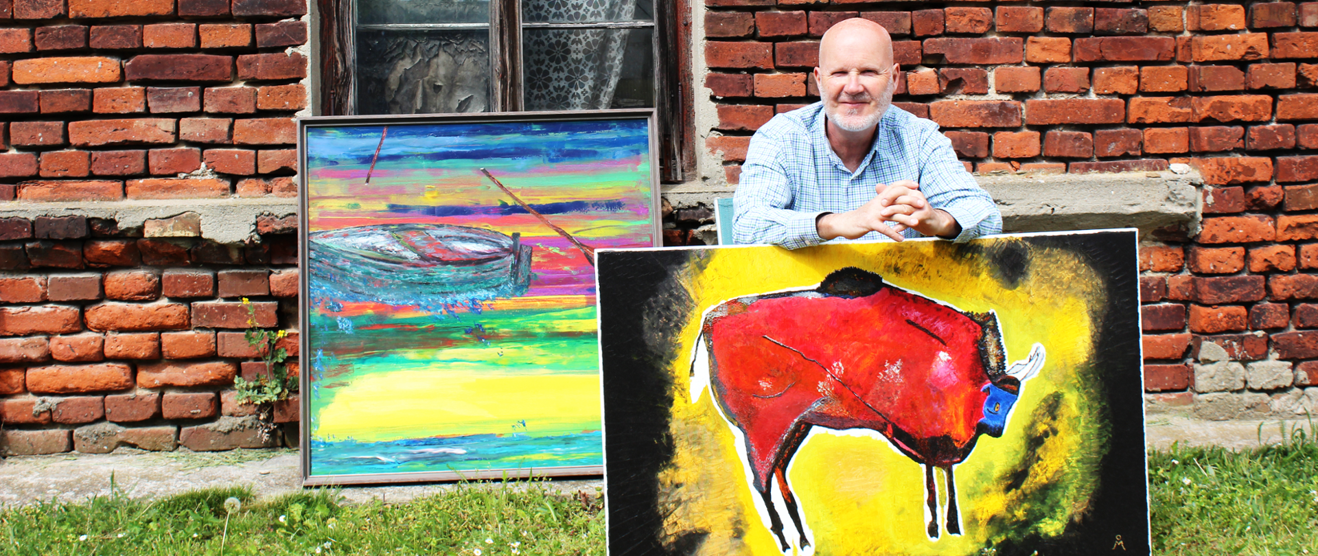 Mężczyzna z siwą brodą, w koszuli w kratę, siedzi na tle ceglanego muru z oknem. Przed nim stoją dwa obrazy: po lewej malowniczy, kolorowy pejzaż z łodzią na wodzie w intensywnych barwach; po prawej ekspresyjny obraz czerwonego byka z niebieską głową na żółto-czarnym tle.