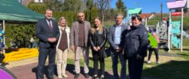 Kilka dorosłych osób pozuje do wspólnego zdjęcia na terenie placu zabaw. W tle po lewej piaskownica z zielonym zadaszeniem, po prawej kolorowy plac zabaw.