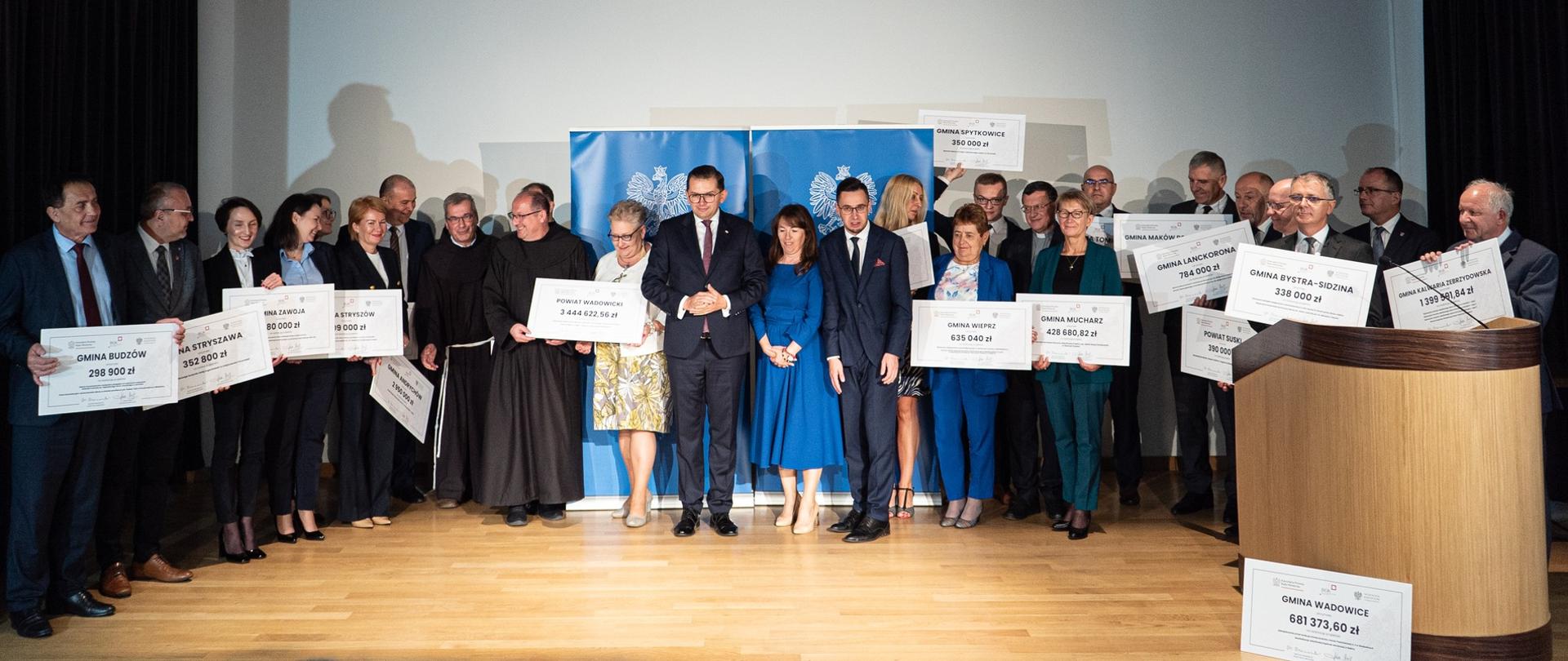 Uczestnicy spotkania pozują do zdjęcia, stojąc w szeregu i trzymając symboliczne czeki potwierdzające dofinansowanie.