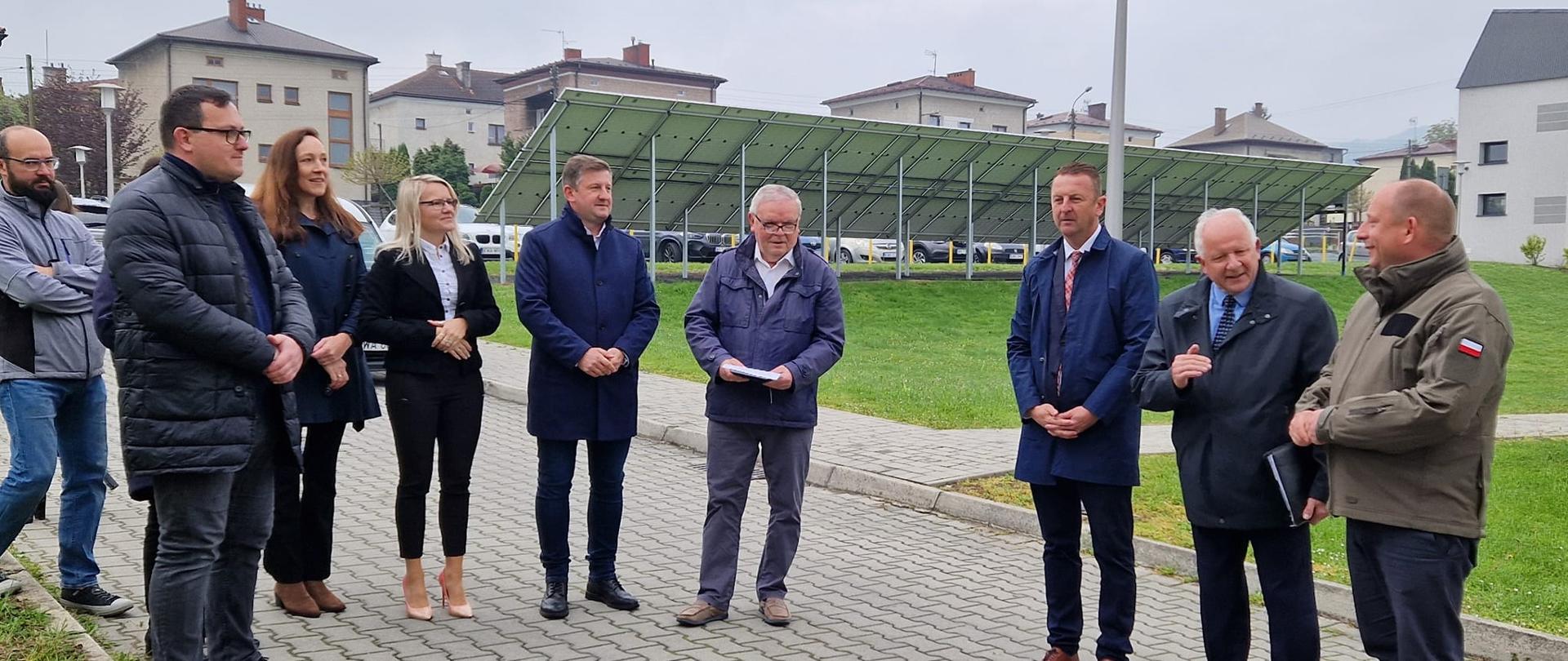 W centrum widać grupę ludzi kobiety i mężczyzn, wśród których stoją Burmistrz Miasta Augustyn Ormanty Wojewoda Małopolski Krzysztof Jan Klęczar Radny Rady Miasta Pracownicy Urzędu Miasta Wykonawca oraz inspektor nadzoru inwestycyjnego wszyscy elegancko ubrani w tle znajdują się budynki i panele fotowoltaiczne 