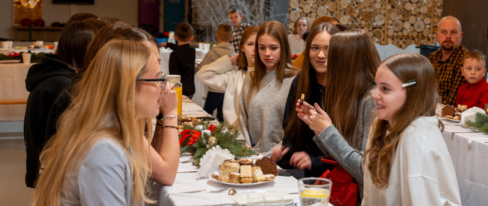 Uczestnicy spotkania opłatkowego zgromadzeni przy długim, świątecznie udekorowanym stole. Młodzież i dorośli prowadzą rozmowy w swobodnej, rodzinnej atmosferze, częstując się ciastem i napojami. Wnętrze sali jest ciepłe i przytulne, z widocznymi dekoracjami świątecznymi oraz naturalnymi elementami wystroju w tle.