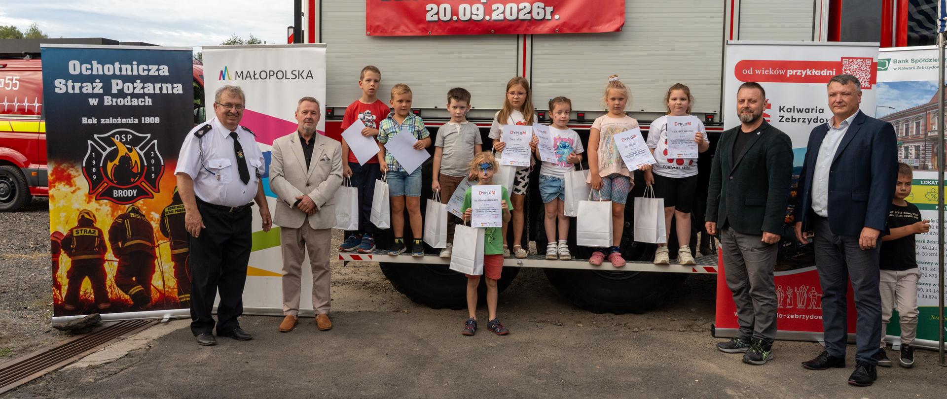 Grupa dzieci z dyplomami i nagrodami stoi przed samochodem strażackim OSP Brody podczas Dnia Bezpieczeństwa 20 września 2026 r. Po bokach stoją przedstawiciele organizatorów i samorządu, a w tle widoczny jest baner wydarzenia oraz roll-upy partnerów.