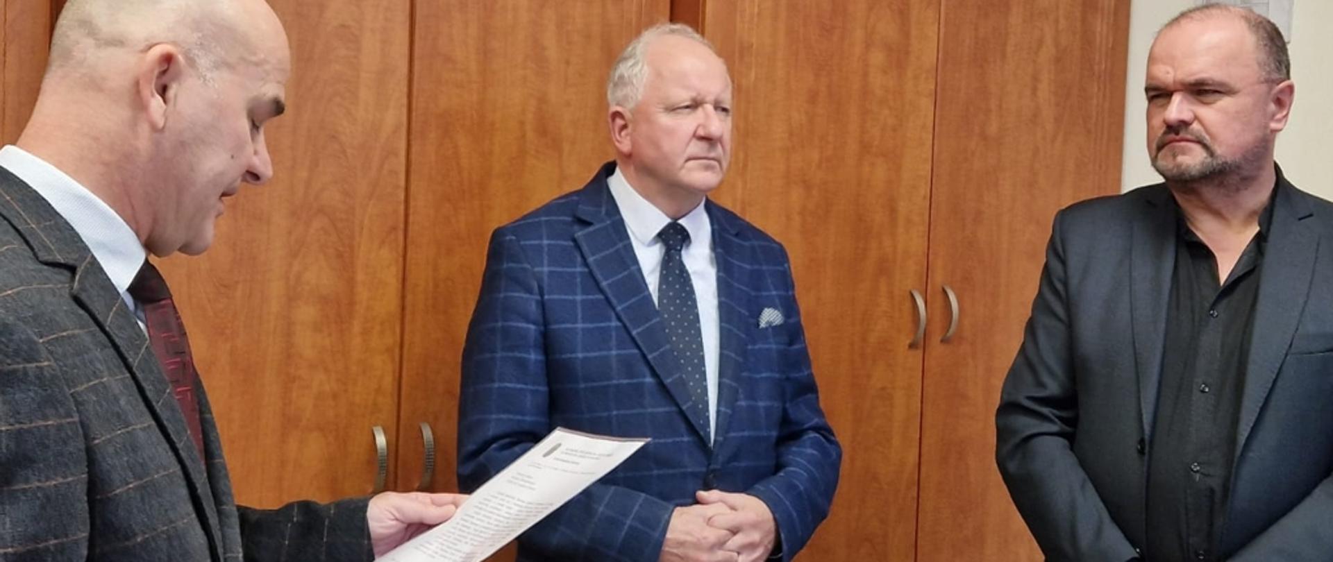 Z lewej strony postać mężczyzny ujęta do połowy, który ma brązowy garnitur, białą koszulę i bordowy krawat. po jego prawej stronie stoi mężczyzna w marynarce granatowej w kratę, jasnej koszuli i krawacie w kropki, na złożone ręce, najbardziej z prawej strony mężczyzna z wąsami czarnymi w ciemniej koszuli i rozpiętej ciemnej marynarce