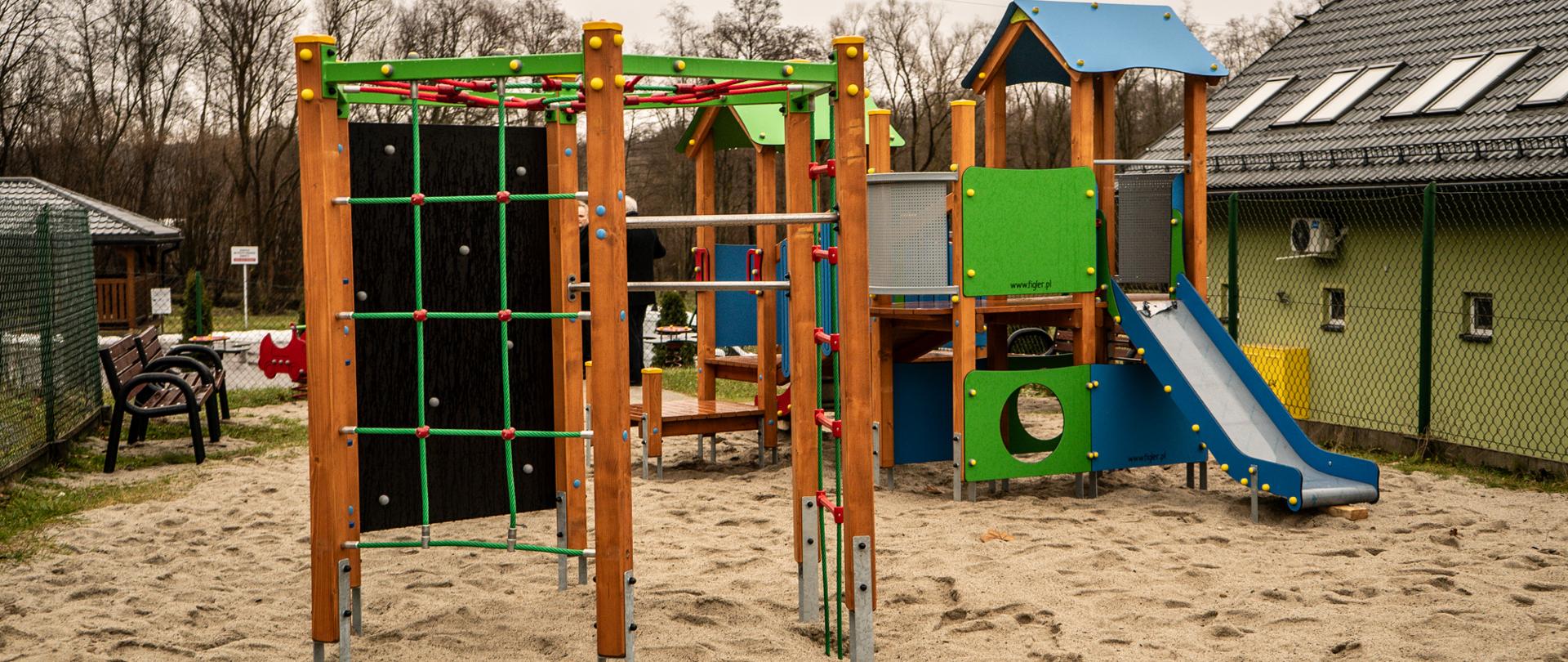 Na zdjęciu przedstawiony został plac zabaw na którym znajdują się sprzęty zabawowe dla dzieci takie jak zjeżdżalnia, ścianka wspinaczkowa,sprzęty te znajdują się na powierzchni z piasku. 