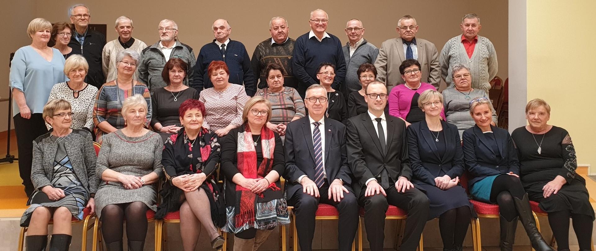 Pamiątkowa fotografia uczestników wigilijki. Zebrani siedzą w dwóch rzędach krzeseł i stoją na scenie. W pierwszym rzędzie Minister Szwed i Wójt Sikorski.