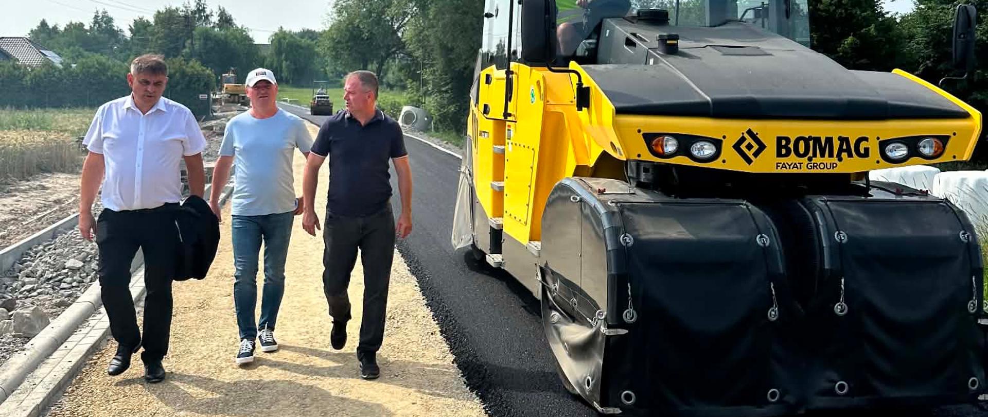 Na zdjęciu trwa przebudowa drogi gminnej. Po prawej stronie widoczny jest duży żółty walec drogowy układający i zagęszczający nową nawierzchnię asfaltową. Po lewej stronie, wzdłuż budowanej jezdni, idą trzy osoby uczestniczące w przeglądzie postępu prac. W tle widać odcinek drogi w trakcie realizacji, ciężki sprzęt budowlany, drzewa oraz zabudowę jednorodzinną. Zdjęcie przedstawia zaawansowany etap inwestycji drogowej w słoneczny dzień.