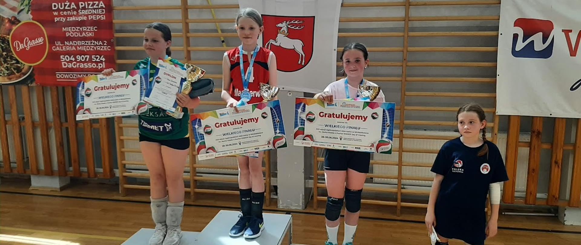 Trzy zawodniczki stoją na podium. Na szyjach mają zawieszone medale, a w rękach trzymają trzymają puchary i dyplomy. Z prawej strony przy podium stoi jeszcze jedna zawodniczka.