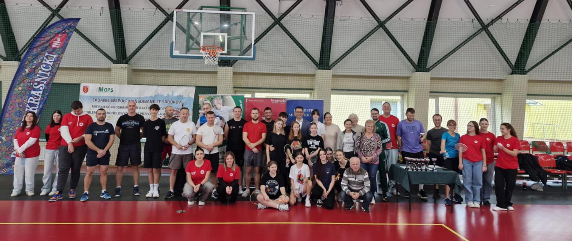 Uczestnicy turnieju stojący w szeregu na hali sportowej. Przed nimi kilka osób kuca lub siedzi na podłodze.