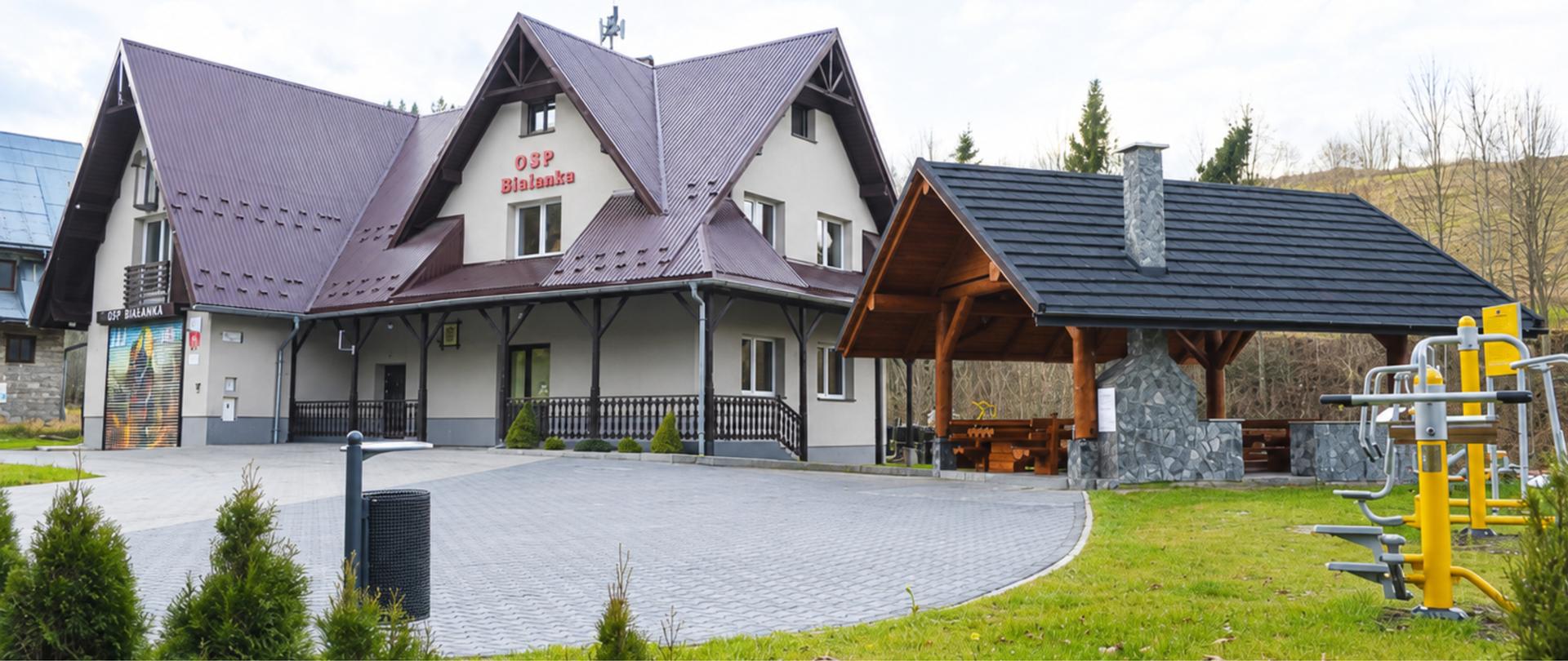 Jasne zdjęcie budynku OSP Bielanka z wielospadowym, ciemnym dachem. Przed budynkiem znajduje się utwardzony plac, a po prawej drewniana wiata z kamiennym kominem oraz zewnętrzna siłownia. Na pierwszym planie widoczny jest trawnik i niewielkie krzewy. W tle znajdują się drzewa oraz łagodne wzgórza pod zachmurzonym, rozjaśnionym niebem.