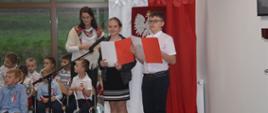 Para konferansjerów - młodzież ze szkoły podstawowej w Sieniawie z teczkami w dłoni w kolorach flagi polski, stoją przed dekoracją w barwach biało-czerwonych. W tle siedzą dzieci z przedszkola oraz Pani opiekunka grupy.