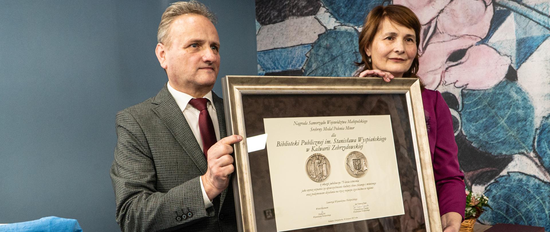 Na ręce Dyrektor Biblioteki Publicznej w Kalwarii Zebrzydowskiej zostaje wręczona Nagroda przez przewodniczącego Komisji Kultury Sejmiku Województwa Małopolskiego Rafała Stuglika. Kobieta i mężczyzna trzymają srebrną ramę stojąc w centrum zdjęcia.