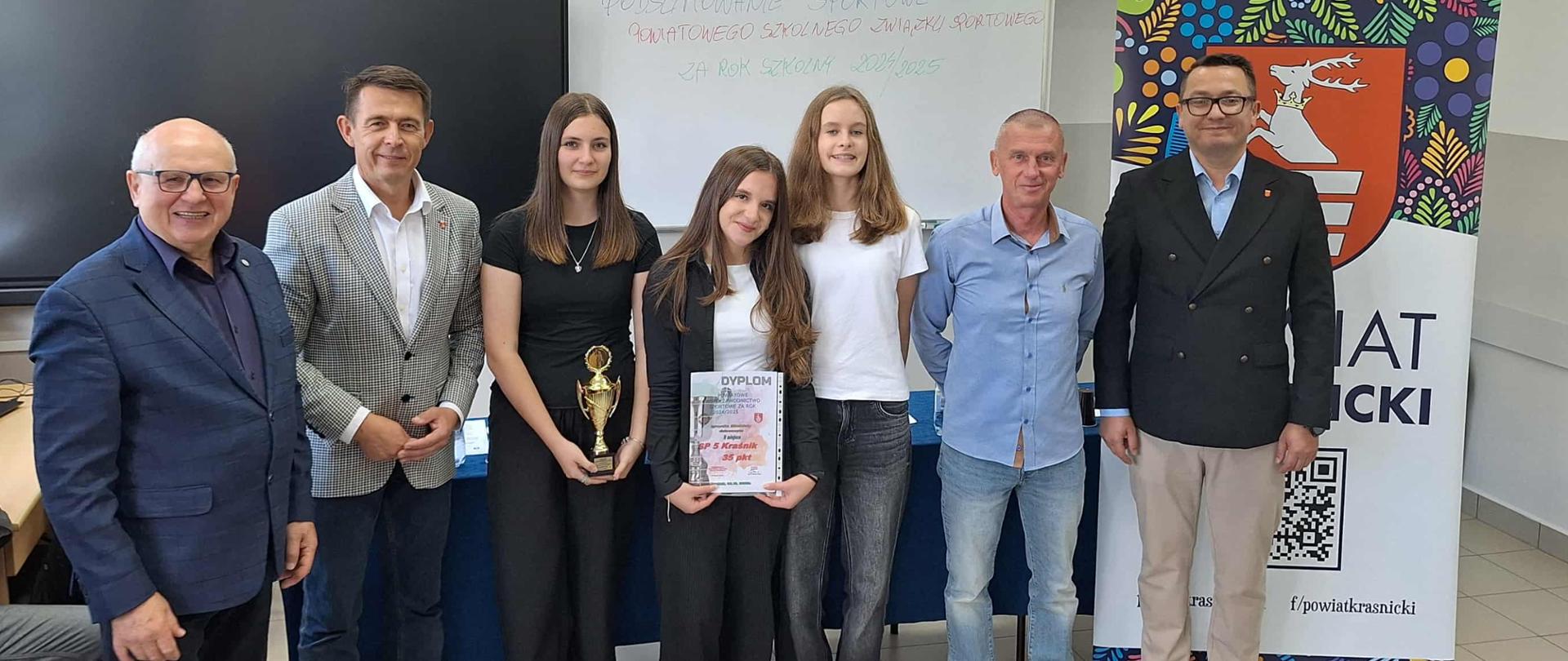 Na tle baneru Powiatu Kraśnickiego i tablic szkolnych stoją od lewej: Przewodniczący Powiatowego Szkolnego Związku Sportowego w Kraśniku Zbigniew Gawdzik, Przewodniczący Rady Powiatu w Kraśniku Jacek Dubiel, trzy uczennice Szkoły Podstawowej nr 5 w Kraśniku z dyplomem i pucharem, nauczyciel Szkoły Podstawowej nr 5 w Kraśniku Jerzy Andrzejczuk oraz sekretarz Miasta Kraśnik Łukasz Skokowski.