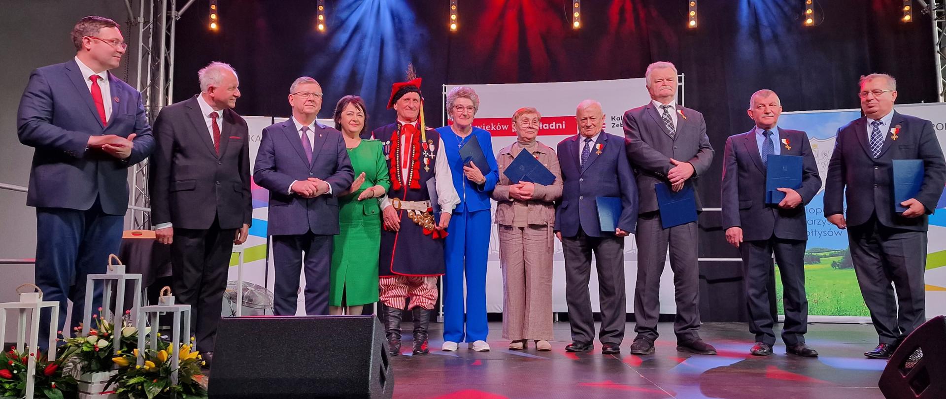 Na scenie jedenaście osób. mężczyźni w garniturach, kobiety w kostiumie. Niektórzy z nich trzymają wręczone im odznaczenia. Nad ich głowami kilka kolorowych reflektorów. 