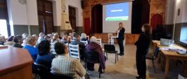Kadr ze spotkania odnośnie renty wdowiej. Prezentacja zagadnienia wyświetlana na rzutniku. Grupa Seniorów siedzi na krzesłach i ogląda prezentacje.