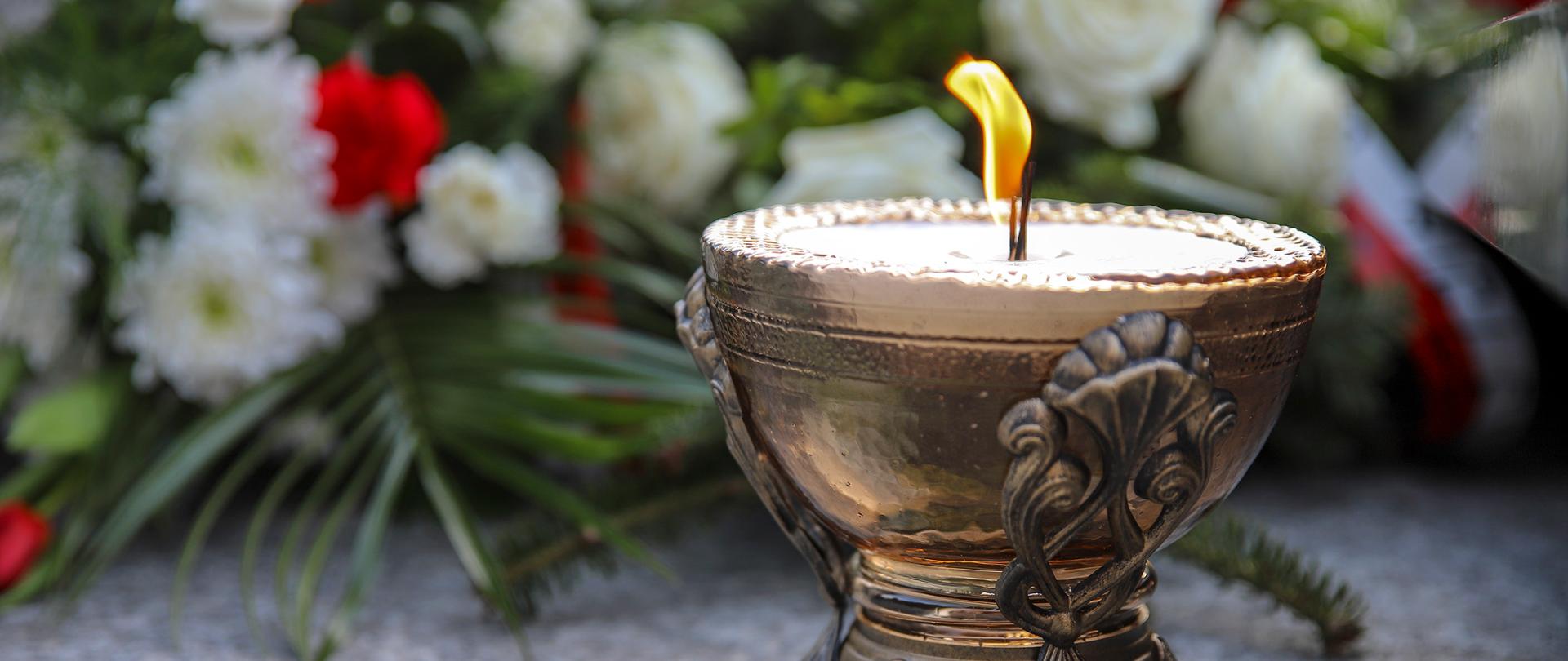 Na zdjęciu z prawej znajduje się zapalony, złoty znicz stojący na granitowej płycie. W tle za nim znajdują się złożone na płycie wiązanki z biało-czerwonych kwiatów.