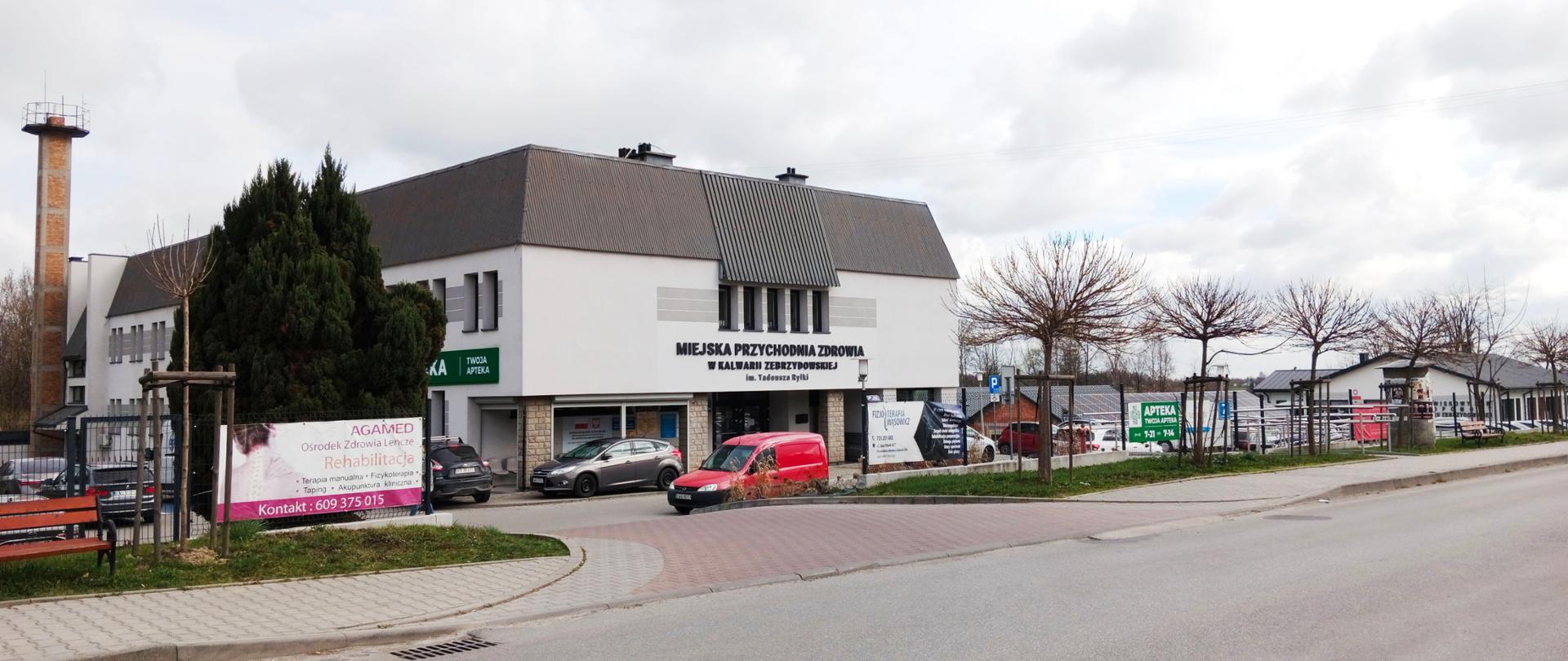 Budynek Miejskiej Przychodni Zdrowia w Kalwarii Zebrzydowskiej widziany z ulicy. Przed wejściem znajdują się zaparkowane samochody, chodnik oraz młode drzewa. Na elewacji widoczny jest napis „Miejska Przychodnia Zdrowia w Kalwarii Zebrzydowskiej”, a obok znajdują się szyldy apteki i gabinetów medycznych. Niebo jest zachmurzone, a całość otacza uporządkowana przestrzeń parkingowa i chodnik przy drodze.
