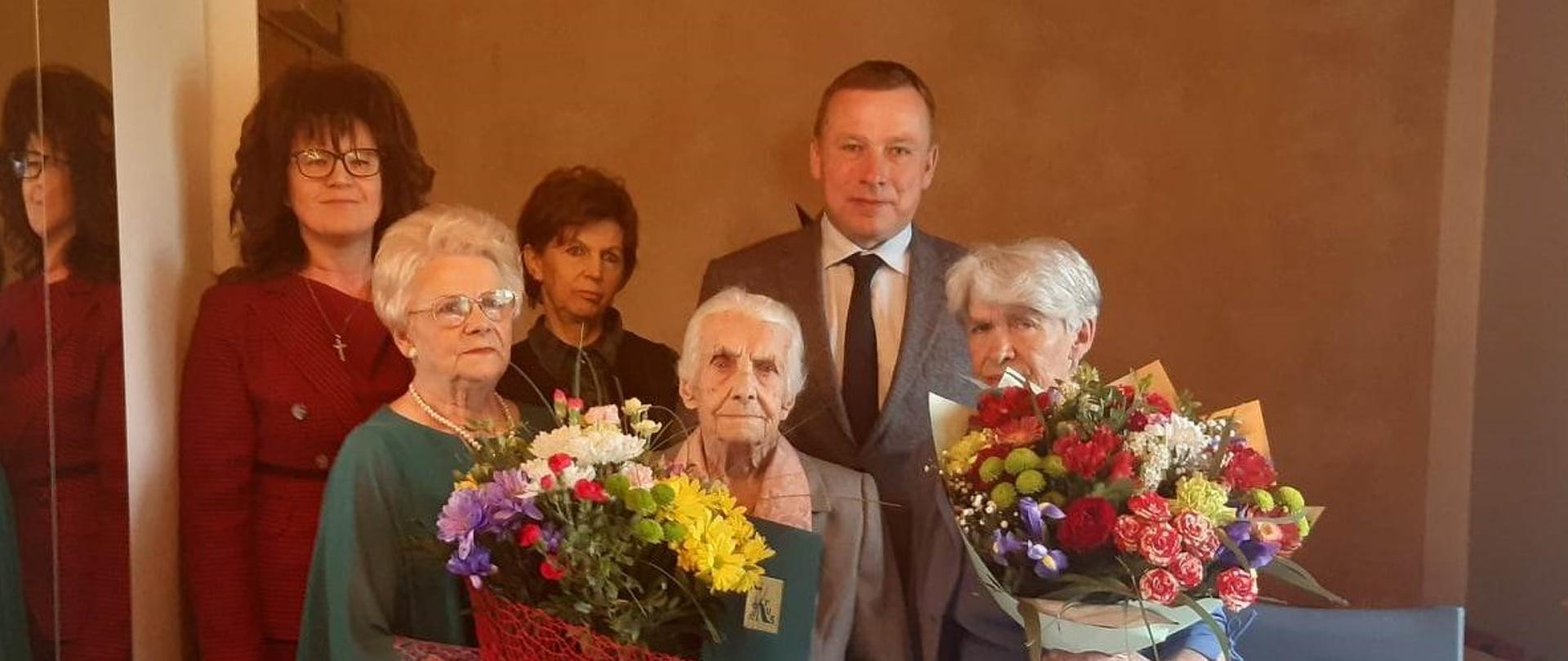 Grupa 6 osób, dwie kobiety po bokach trzymają bukiety kwiatów, kobieta w środku zieloną teczkę