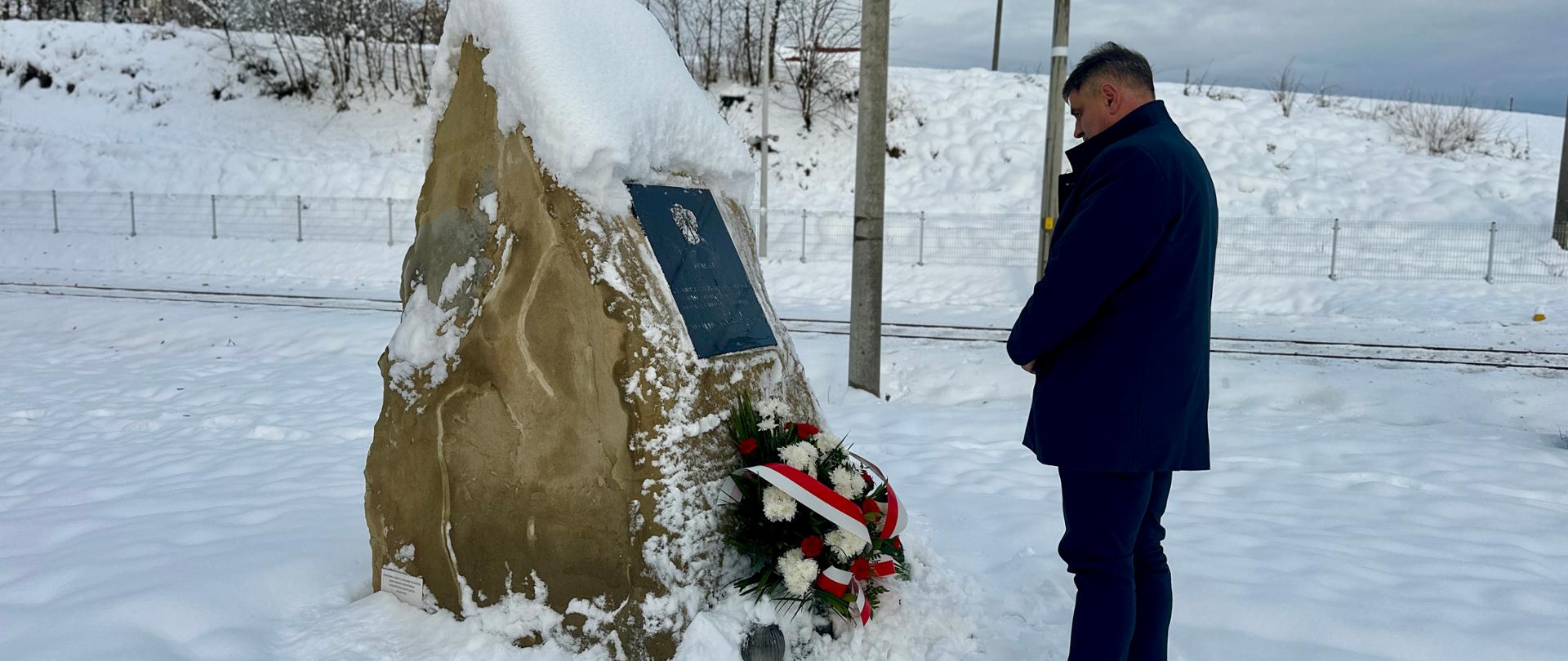 Burmistrz Kalwarii Zebrzydowskiej stoi przed ośnieżonym obeliskiem upamiętniającym ofiary katastrofy kolejowej w Barwałdzie Średnim. Na pomniku widoczna jest tablica pamiątkowa i złożony biało-czerwony wieniec. W tle widać zasypane śniegiem torowisko oraz stoki wzgórza z drzewami.