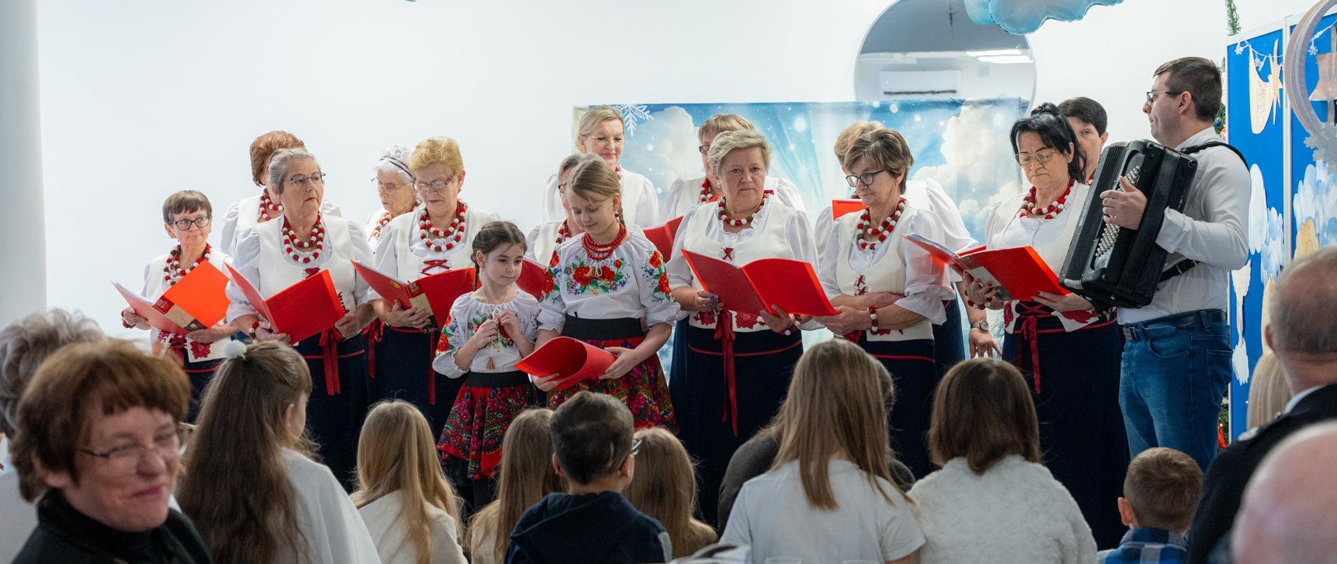 Grupa kobiet i dzieci w tradycyjnych strojach ludowych śpiewa kolędy, trzymając czerwone śpiewniki; z boku akompaniuje mężczyzna grający na akordeonie, przed nimi zgromadzona publiczność siedząca przy stołach w jasnej sali udekorowanej zimowymi motywami i niebieskimi balonami.