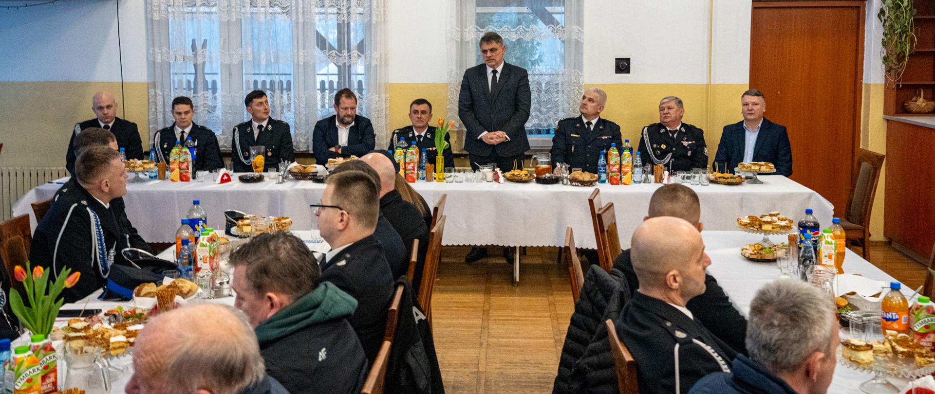 Sala remizy strażackiej wypełniona uczestnikami walnego zebrania OSP. Przy długim, nakrytym białym obrusem stole siedzą zaproszeni goście i przedstawiciele służb w mundurach. W centralnej części stoi mężczyzna w garniturze, przemawiający do zebranych. Na stołach widoczne są napoje, poczęstunek oraz dekoracje kwiatowe, a w tle okna z firanami i świąteczne oświetlenie.