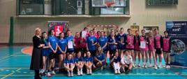 Zawodniczki biorące udział w turnieju stojące w tylnych rzędach i siedzące na podłodze w rzędzie pierwszym na środku hali sportowej z dyplomami i pucharami w rękach. Od lewej strony stoi Wicedyrektor Zespołu Szkół nr 3 w Kraśniku Monika Gozdalska.