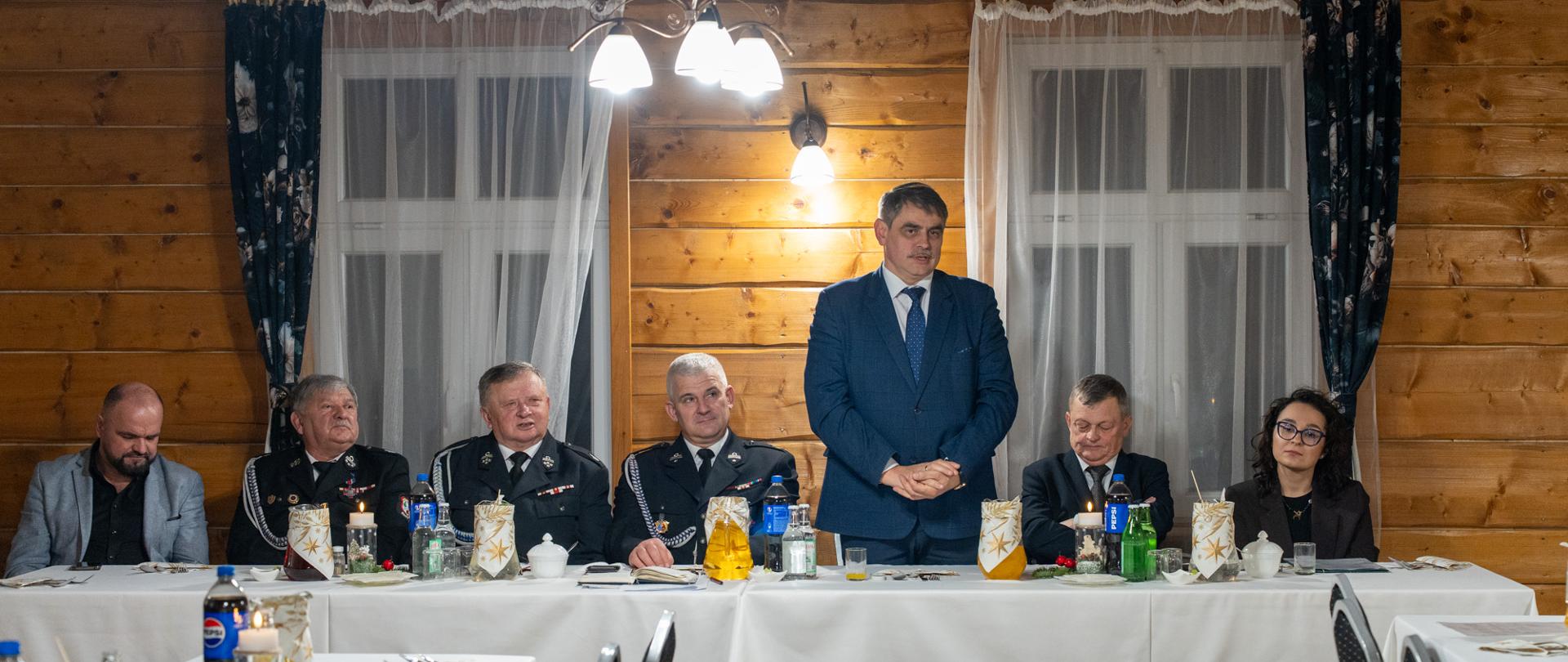 Uczestnicy zebrania i spotkania wigilijnego siedzą przy długim stole w drewnianej sali. W centrum mężczyzna w garniturze stoi i przemawia do zgromadzonych. Obok niego siedzą przedstawiciele Ochotniczej Straży Pożarnej w mundurach oraz zaproszeni goście. Stół jest nakryty białym obrusem, z dekoracjami świątecznymi, świecami i napojami, co podkreśla oficjalno-uroczysty charakter wydarzenia.