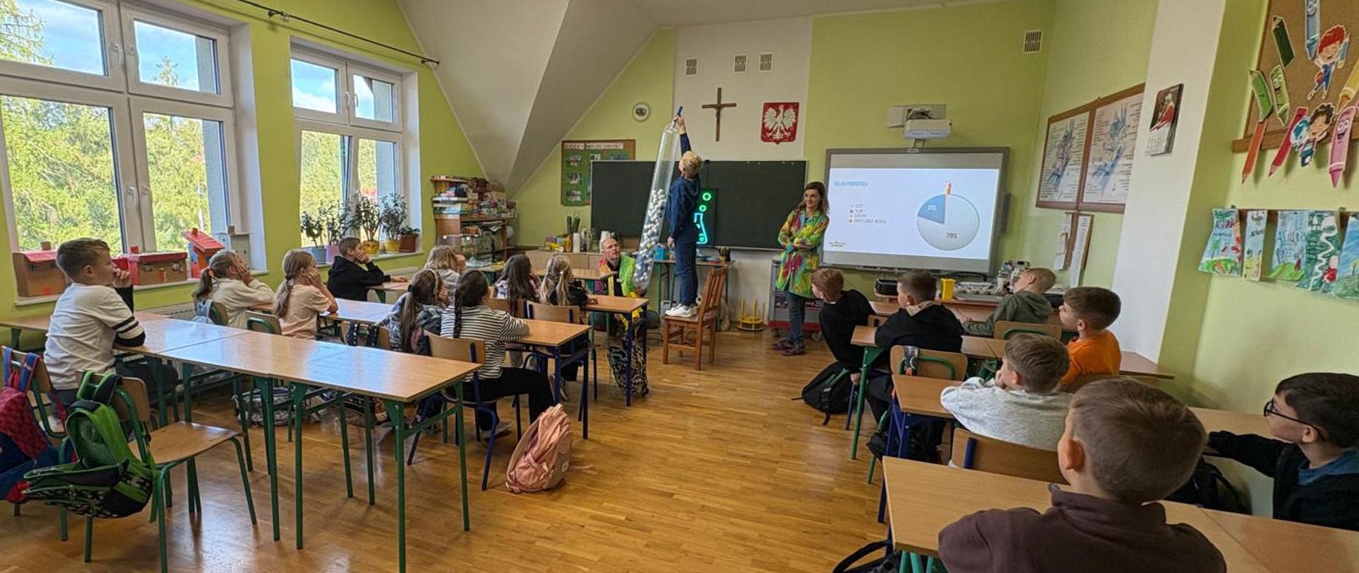 Sala lekcyjna z uczniami siedzącymi w ławkach, pod tablicą stoją prowadząca zajęcia i uczeń wykonujący doświadczenie
