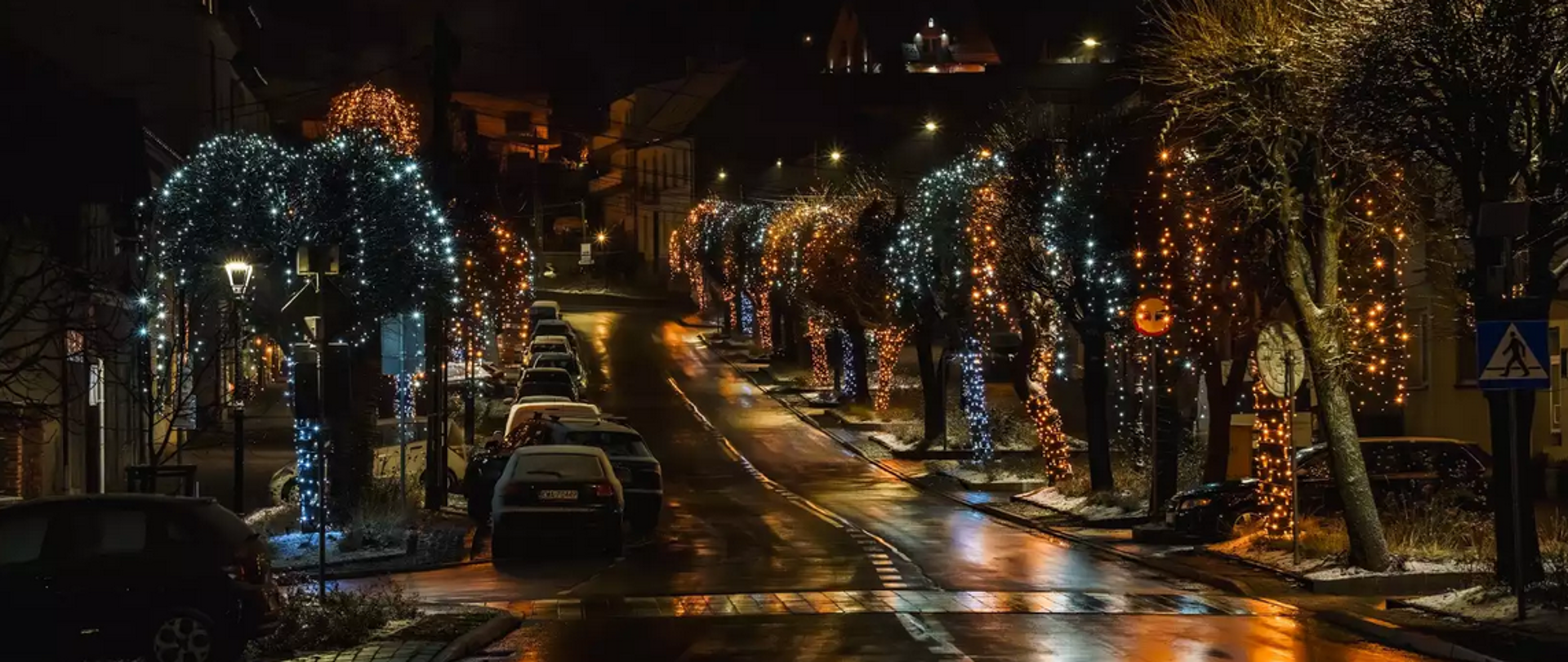 Nocny widok na ulicę w Kalwarii Zebrzydowskiej ozdobioną świątecznymi iluminacjami. Drzewa wzdłuż jezdni oplecione są białymi i złotymi światłami, które odbijają się w mokrym asfalcie, tworząc efektowne refleksy. Po obu stronach ulicy zaparkowane są samochody, a w tle widoczna jest zabudowa miasta. Całość tworzy nastrojowy, zimowy klimat świątecznego wieczoru.