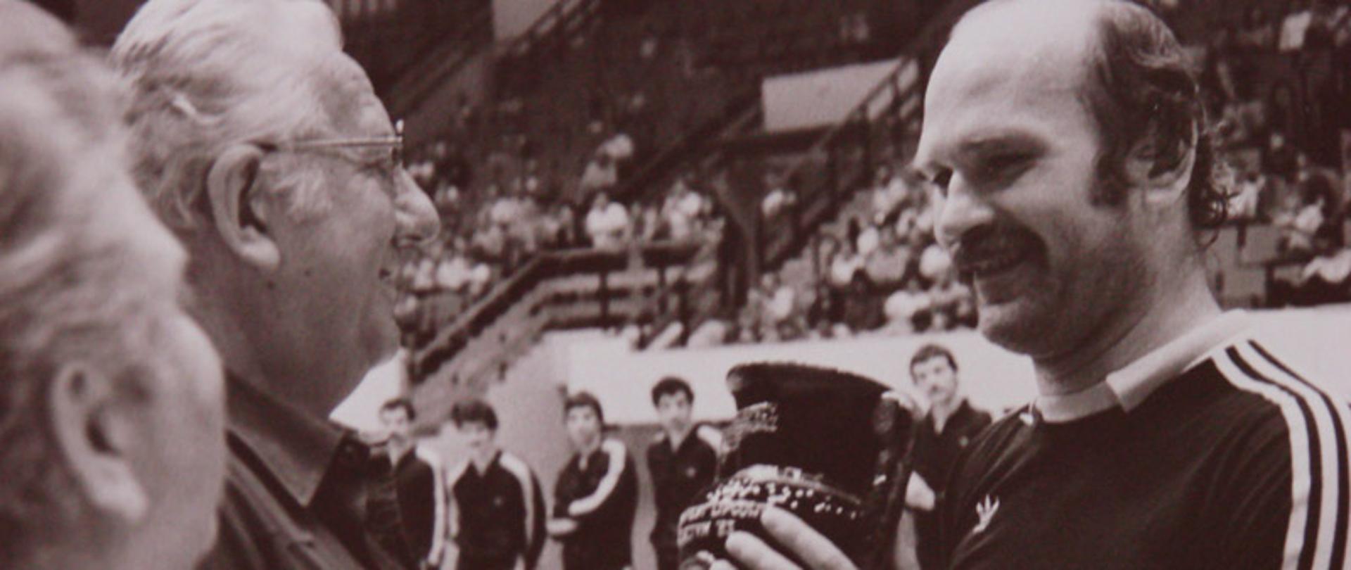 Czarno-białe zdjęcie przedstawiające uśmiechniętego Andrzeja Szymczaka w stroju sportowym, odbierającego puchar w hali sportowej z rąk mężczyzny w okularach.
