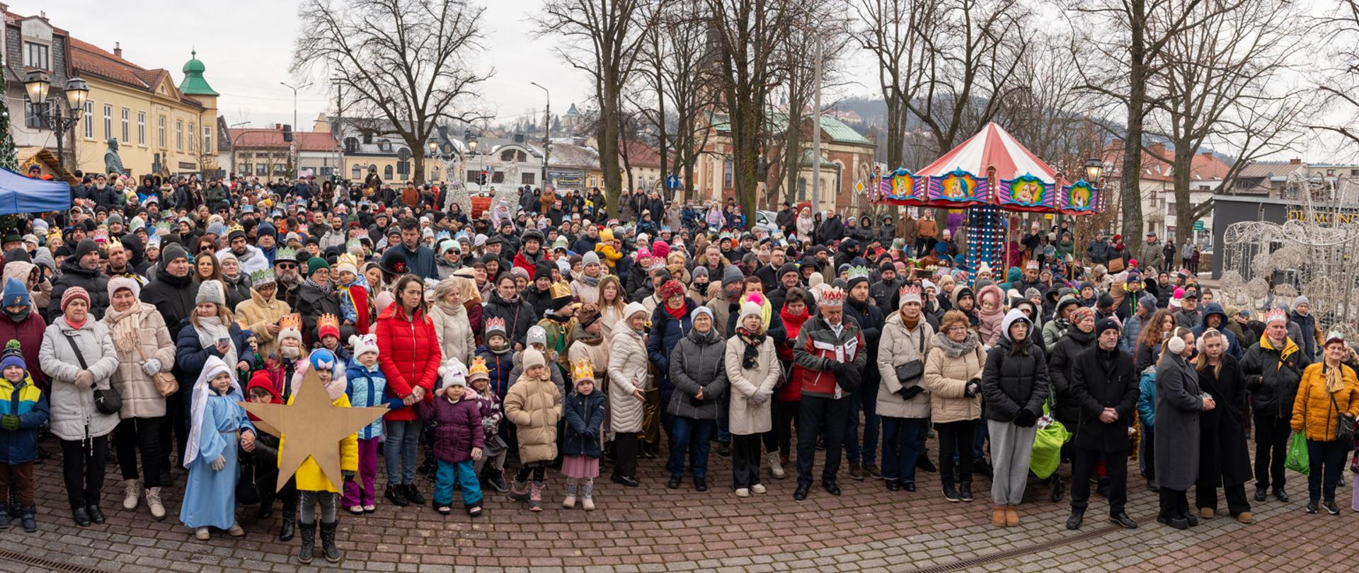 Panoramiczne zdjęcie Rynku w Kalwarii Zebrzydowskiej wypełnionego licznymi uczestnikami Orszaku Trzech Króli. Wśród zgromadzonych widoczne są rodziny z dziećmi, osoby w zimowych płaszczach, wielu uczestników w kolorowych papierowych koronach oraz dzieci przebrane w stroje jasełkowe. W tle zabudowa rynku, drzewa bez liści, świąteczne dekoracje i karuzela. Atmosfera uroczysta i radosna.