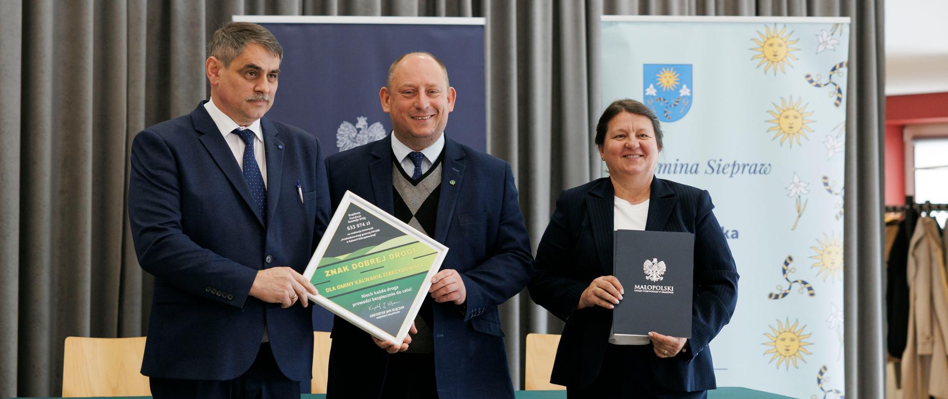 Na zdjęciu trzech przedstawicieli administracji publicznej podczas uroczystości podpisania umów o dofinansowanie. W centrum Burmistrz Kalwarii Zebrzydowskiej wraz z Wojewodą Małopolskim prezentują dokument potwierdzający przyznane środki, obok stoi Skarbnik Gminy z teczką z logo Małopolski. W tle widoczne banery instytucjonalne oraz kurtyna, podkreślające oficjalny charakter wydarzenia. 