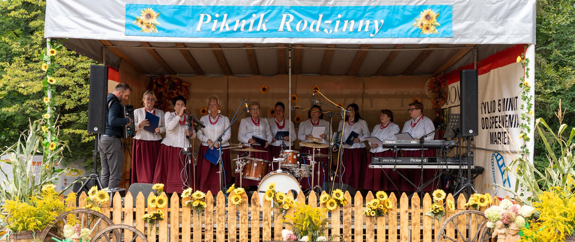 Występ zespołu podczas Pikniku Rodzinnego w Barwałdzie Górnym. Scena udekorowana jest słonecznikami, kwiatami, drewnianymi kołami i płotkiem w rustykalnym stylu.