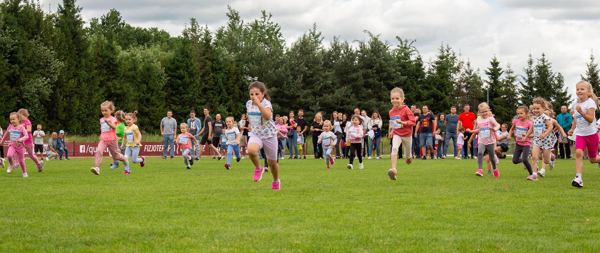 Grupa małych dzieci biegnie po trawie na otwartym boisku, uczestnicząc w biegu lub zawodach sportowych. Na pierwszym planie widać kilka dziewczynek w kolorowych ubraniach, skupionych na biegu. W tle stoją dorośli – prawdopodobnie rodzice i opiekunowie – obserwujący wydarzenie. Za nimi widoczna jest linia drzew i lekko zachmurzone niebo.