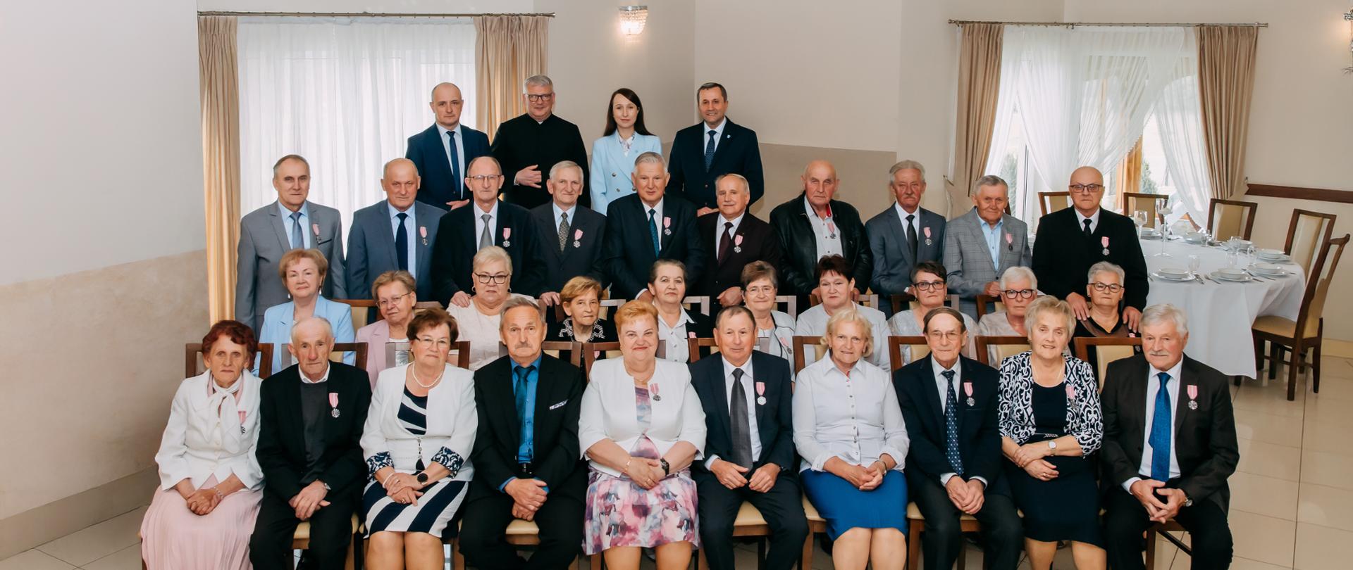 Wszyscy jubilaci wspólnie pozują do zdjęcia w trzech rzędach, ostatni czwarty rząd to Wójt Gminy, Kierownik USC, Ksiądz Proboszcz z Parafii z Raby Wyżnej oraz Przewodniczący Gminy Raba Wyżna. 