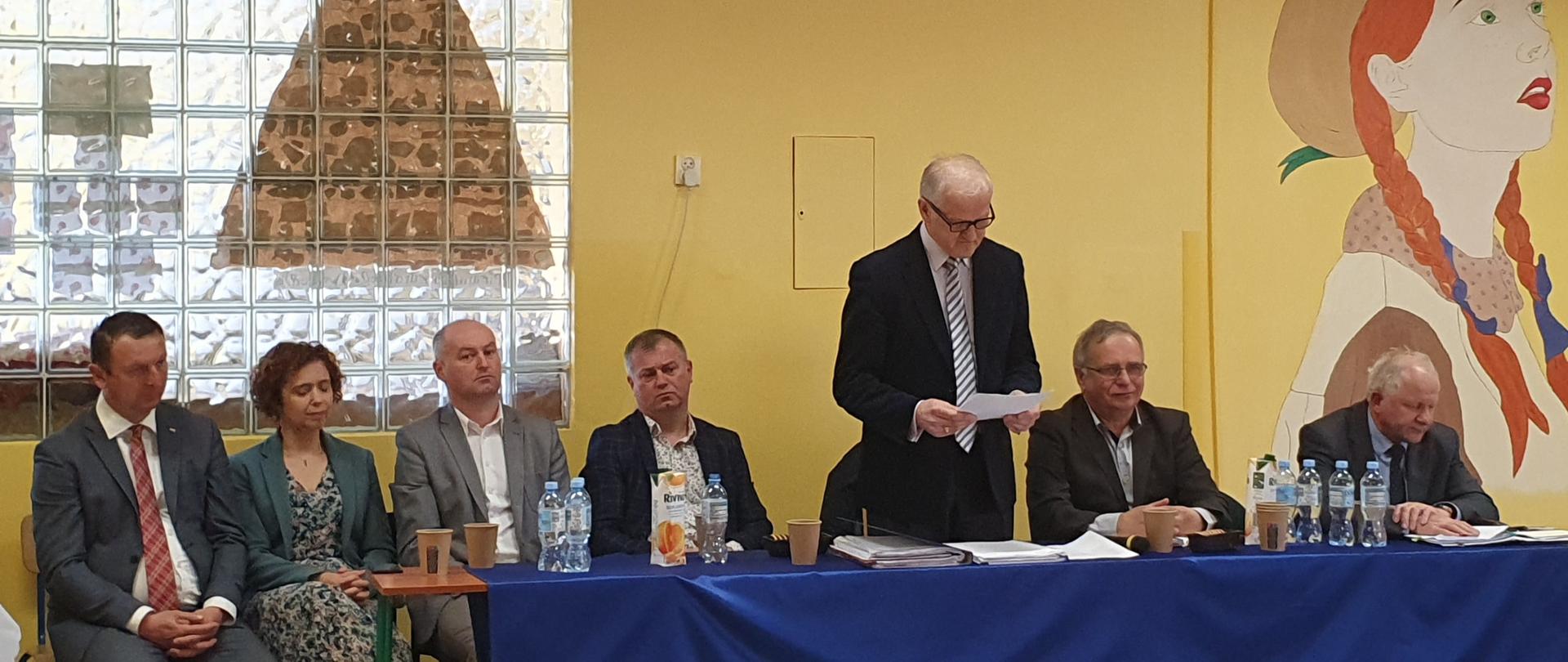 Na szkolnym korytarzu połączone stoliki nakryte granatowym suknem. Przy stole siedzą radni rady miejskiej, dyrektorka szkoły, burmistrz, radny powiatowy. Pośrodku nich na stojąco przemawia sołtys, czytając z kartki trzymanej obiema rękami.