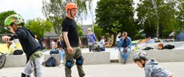 Instruktor w kasku i ochraniaczach na kolanach. Dwóch chłopców jedzie na deskorolkach. W tle skatepark i grupka przyglądających się ludzi.