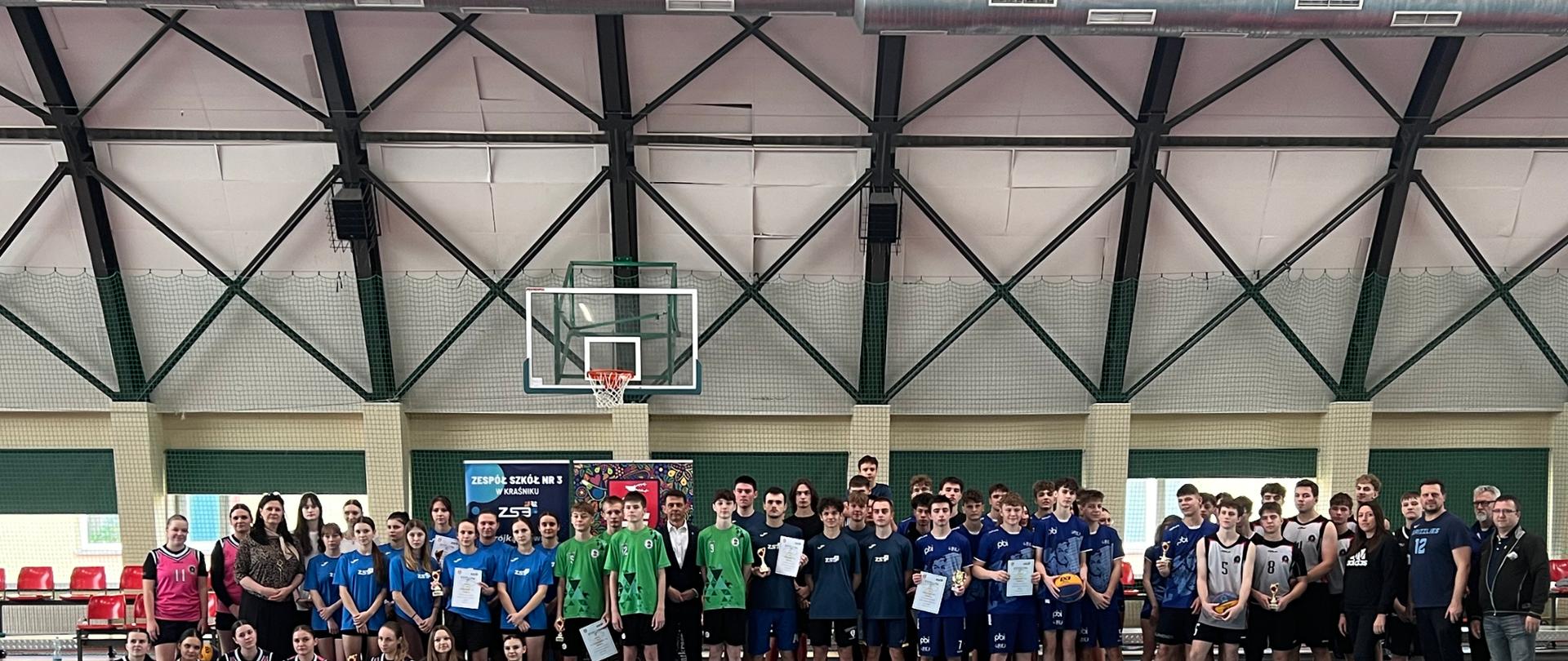 Uczestnicy turnieju stojący na hali sportowej z dyplomami i pucharami w rękach