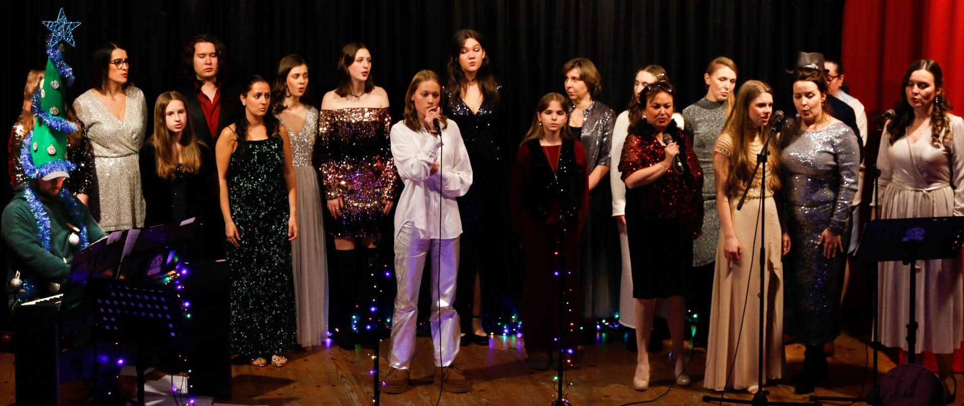 Grupa osób elegancko ubranych na scenie śpiewa wspólnie kolędę, od lewej strony pianista który akompaniuje . Scena udekorowana jest światełkami świątecznymi tło kurtyna czarna.