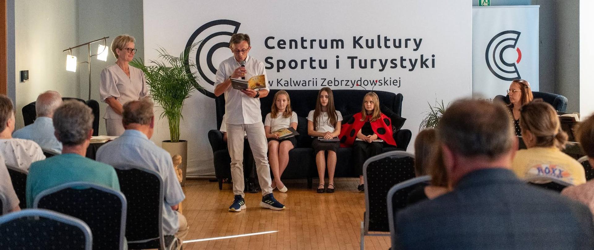 Na sali Centrum Kultury, Sportu i Turystyki w Kalwarii Zebrzydowskiej odbywa się spotkanie autorskie. Na środku stoi mężczyzna czytający fragment książki do mikrofonu, obok niego stoi kobieta. Za nimi na scenie siedzą trzy dziewczynki oraz prowadząca wydarzenie. Przed sceną zgromadzona jest publiczność siedząca na krzesłach. W tle widoczne jest duże logo i nazwa Centrum Kultury, Sportu i Turystyki w Kalwarii Zebrzydowskiej.