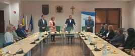 Grupa dorosłych siedząca przy stole. Na stole znajdują się butelki z wodą, szklanki oraz talerzyki z ciastem. W tle sala konferencyjna z flagami: Gminy Raba Wyżna, Polski, UE po lewej stronie. Na środku Herb Gminy Raba Wyżna, Godło Polski, Krzyż. Po prawej stronie baner z logiem Gminy.