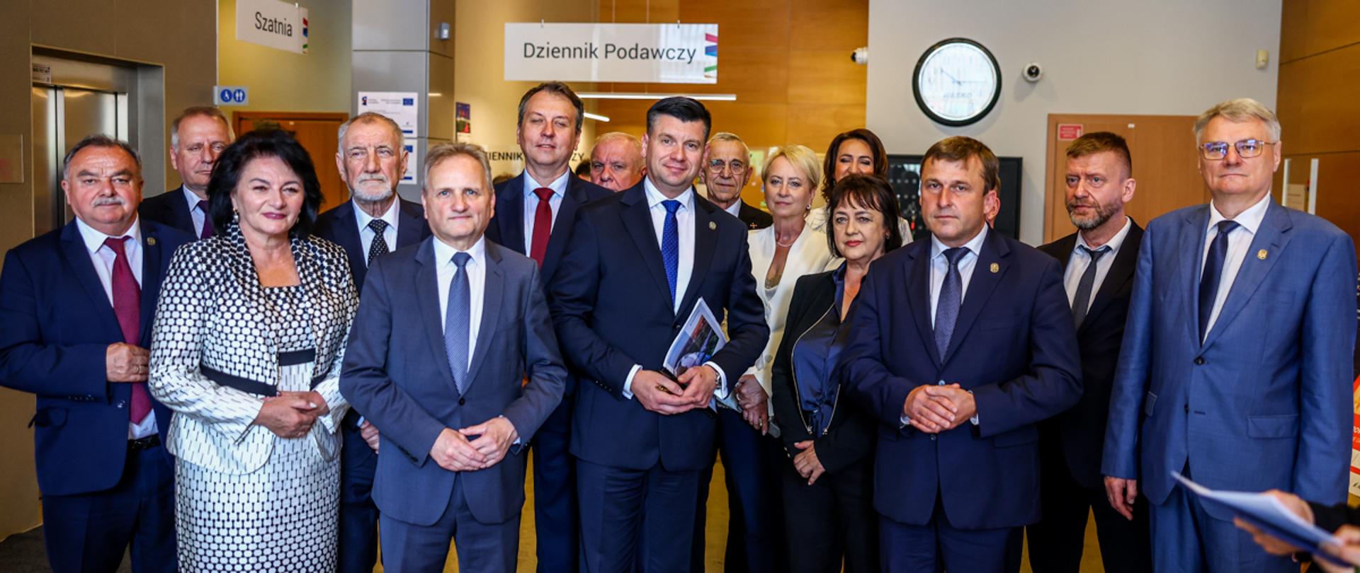 Grupa kilkunastu przedstawicieli samorządów oraz administracji publicznej pozuje do wspólnego zdjęcia wewnątrz nowoczesnego budynku urzędu. W centrum stoją mężczyźni w garniturach, a wokół nich pozostali uczestnicy wydarzenia, w tym kobiety i mężczyźni ubrani w stroje formalne. W tle widoczne są tablice informacyjne z napisami „Dziennik Podawczy” i „Szatnia”, zegar ścienny oraz hol urzędu z drewnianym wykończeniem. Wszyscy uczestnicy zwróceni są w stronę aparatu i pozują do pamiątkowej fotografii.