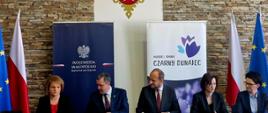 Pięć osób ubranych na galowo siedzących za stołem podpisują umowę, od lewej kobieta, dwóch mężczyzn, dwie kobiety, w tle roll-up, po bokach w tle flaga europejska i flaga Polski