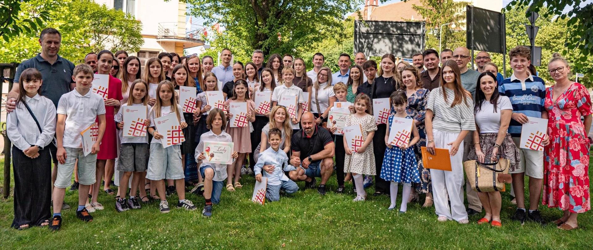 Grupowe zdjęcie uczestników i organizatorów uroczystości wręczenia stypendiów artystycznych. Na trawniku przed budynkiem Urzędu Miejskiego stoi kilkadziesiąt osób - dzieci, młodzież i dorośli. Wiele osób trzyma dyplomy oraz teczki ze stypendiami. W tle widoczne są drzewa oraz zabudowa centrum Konstantynowa Łódzkiego. Uczestnicy pozują do wspólnego pamiątkowego zdjęcia po zakończeniu uroczystości.