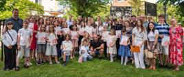 Grupowe zdjęcie uczestników i organizatorów uroczystości wręczenia stypendiów artystycznych. Na trawniku przed budynkiem Urzędu Miejskiego stoi kilkadziesiąt osób - dzieci, młodzież i dorośli. Wiele osób trzyma dyplomy oraz teczki ze stypendiami. W tle widoczne są drzewa oraz zabudowa centrum Konstantynowa Łódzkiego. Uczestnicy pozują do wspólnego pamiątkowego zdjęcia po zakończeniu uroczystości.