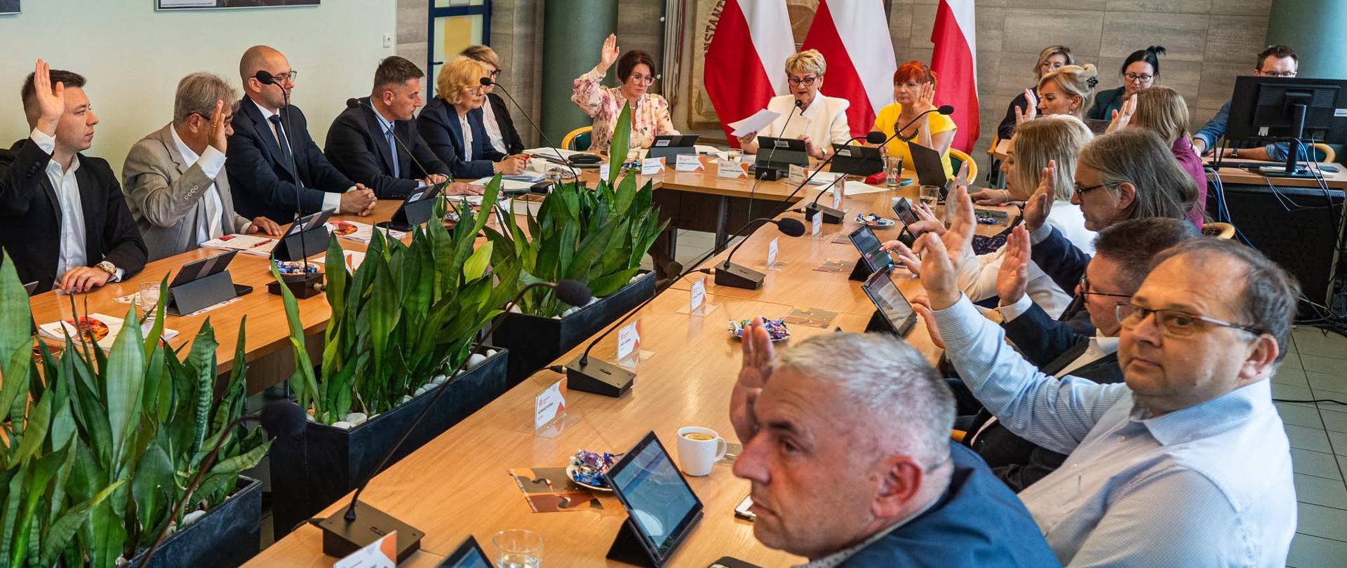 Posiedzenie Rady Miejskiej w Konstantynowie Łódzkim. Radni siedzą przy długich stołach konferencyjnych wyposażonych w mikrofony i tablety. W trakcie głosowania większość uczestników podnosi ręce. Na środku sali znajdują się donice z zielonymi roślinami, a w tle widoczne są biało-czerwone flagi oraz prezydium obrad.